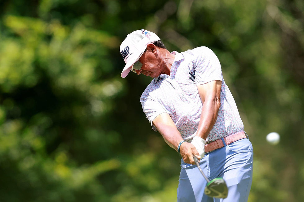 Golfer in white cap and patterned shirt swings a club on a green course; blurred trees behind, focused action, TMP on cap