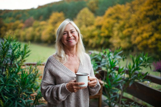 Smiling blonde woman holding a white mug on a porch, with autumn trees and greenery in the background.