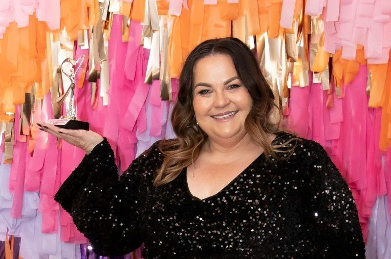 Smiling woman in black sequined dress holds a trophy. Colorful paper backdrop in pink, orange, and gold creates a festive atmosphere.