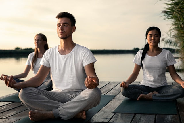 Three people meditate on yoga mats on a wooden dock by a calm lake at sunset.