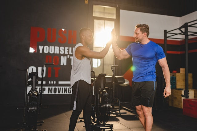 Two men high-five in a gym, with a wall slogan: PUSH YOURSELF DON'T QUIT. Warm, upbeat workout scene.