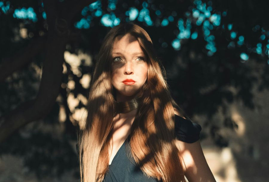 A young woman with long, straight hair stands under dappled sunlight, with shadows of leaves cast across her face and body.