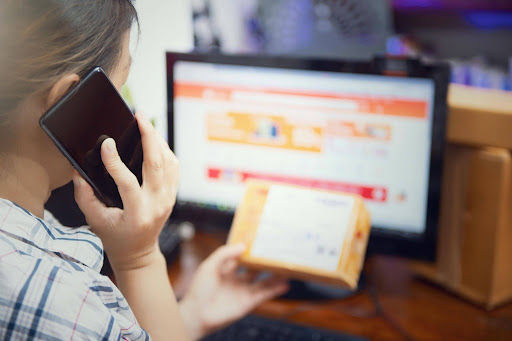 Person on phone holds package, facing a blurred computer screen, suggesting online shopping. Plaid shirt, warm lighting, busy mood.