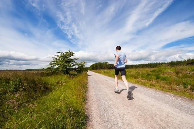 Man running on a gravel path through lush countryside, under a blue sky with scattered clouds. Vibrant green grass lines the path.