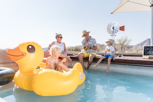 Family and kids relax by a desert pool on a giant yellow duck float, with a windmill and chalkboard sign in the background.