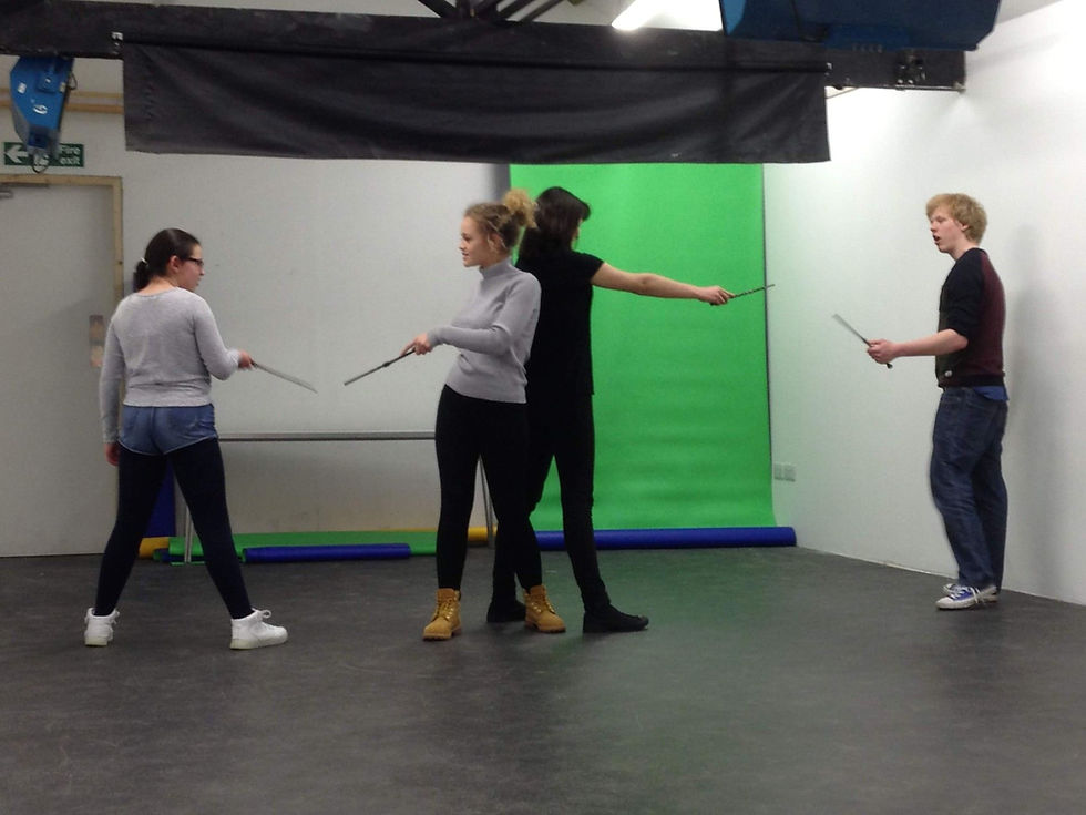 Four young adults practice stage combat with swords in a studio, standing before a green screen and white walls.