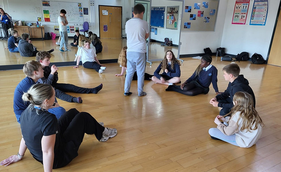 Students sit in a circle on a polished studio floor while a teacher stands in the center; mirrors, posters, and bags line the walls.
