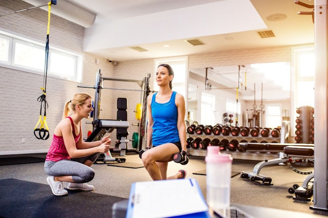 Two women in a gym; one in a blue shirt doing lunges with dumbbells, the other in pink coaching beside. Bright, clean setting with gym equipment.