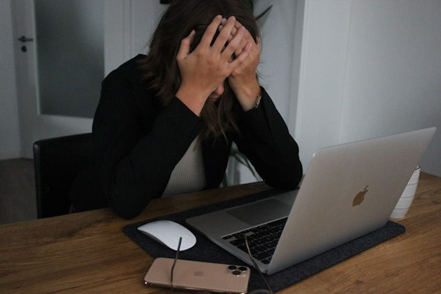 A tired woman sits at a desk with her head in her hands, looking overwhelmed in front of an open laptop.