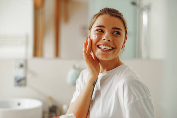 Woman smiling in a bathroom, applying cream to her cheek. She wears a white shirt. The background shows a sink and soft, neutral tones.