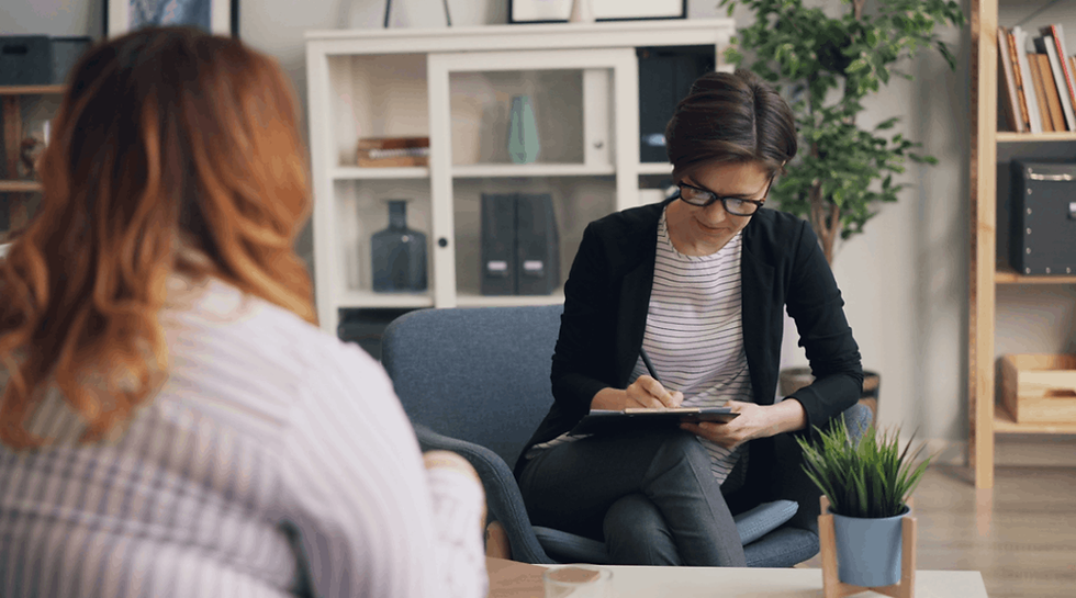 Therapist in glasses takes notes while listening to a woman in a bright office with shelves, plants, and a calm mood.