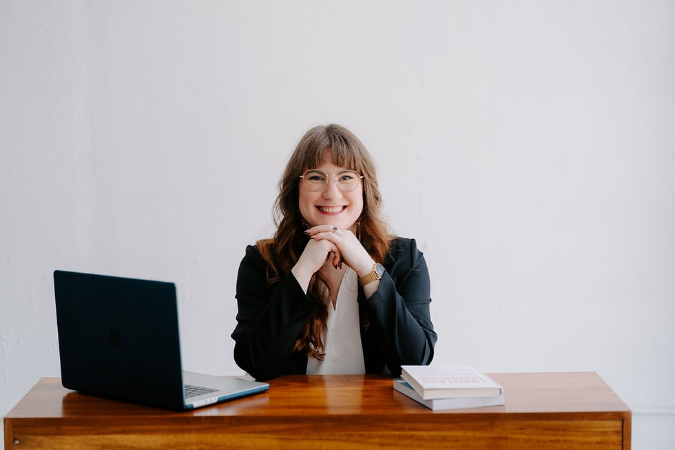 Smiling woman in a black blazer sits at a wooden desk with a laptop and a book. White background, glasses, and watch visible.