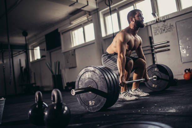 Man lifting barbell in a gym, surrounded by kettlebells. He's shirtless, focused, with sunlight streaming through windows. Determined mood.