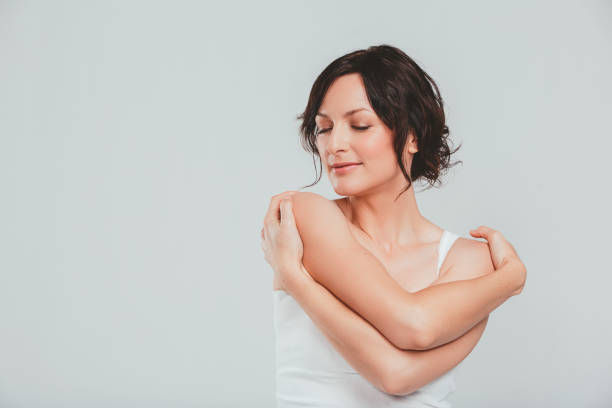 Woman with short dark hair in a white tank top hugs herself, eyes closed, smiling peacefully against a plain light gray background.