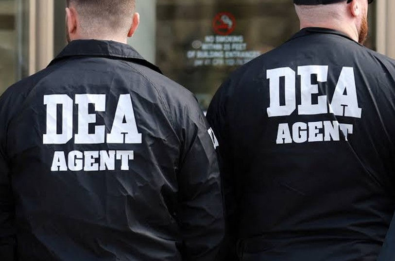 Two DEA agents seen from behind in black jackets with large white DEA AGENT text, standing outside a building.