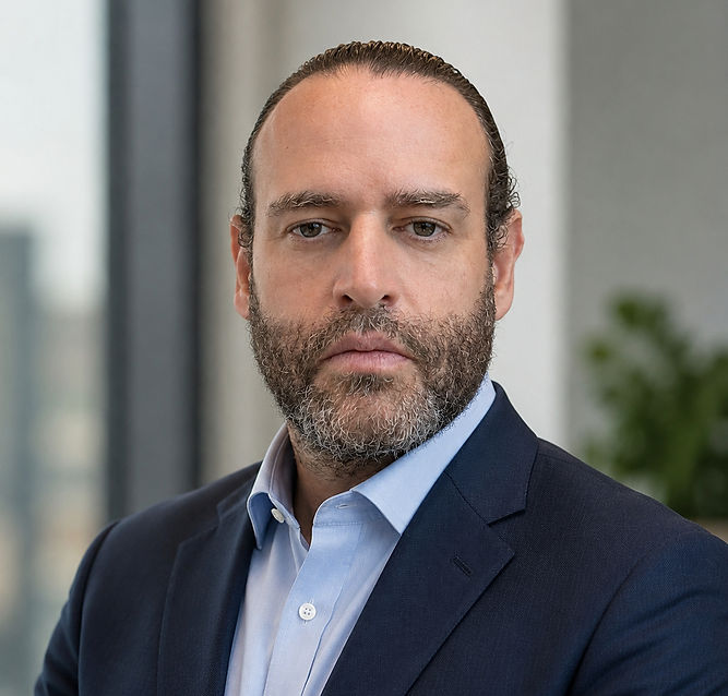 Serious man in a navy suit and light blue shirt, posing in a bright office with blurred windows and a plant behind him.