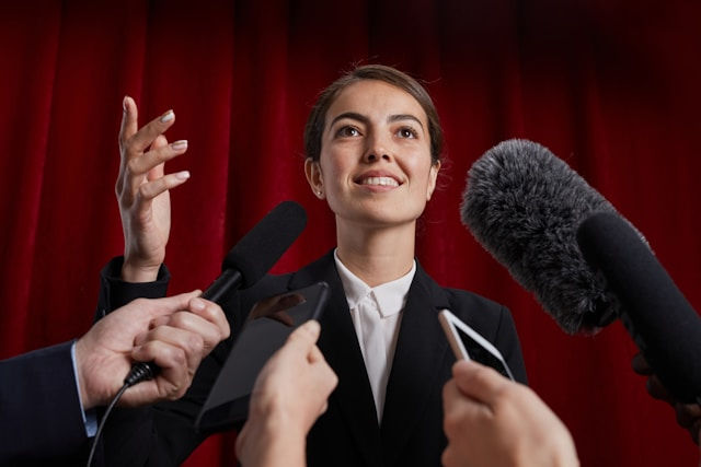 Woman in a suit speaking to multiple microphones, red curtain backdrop. She appears confident and engaged.