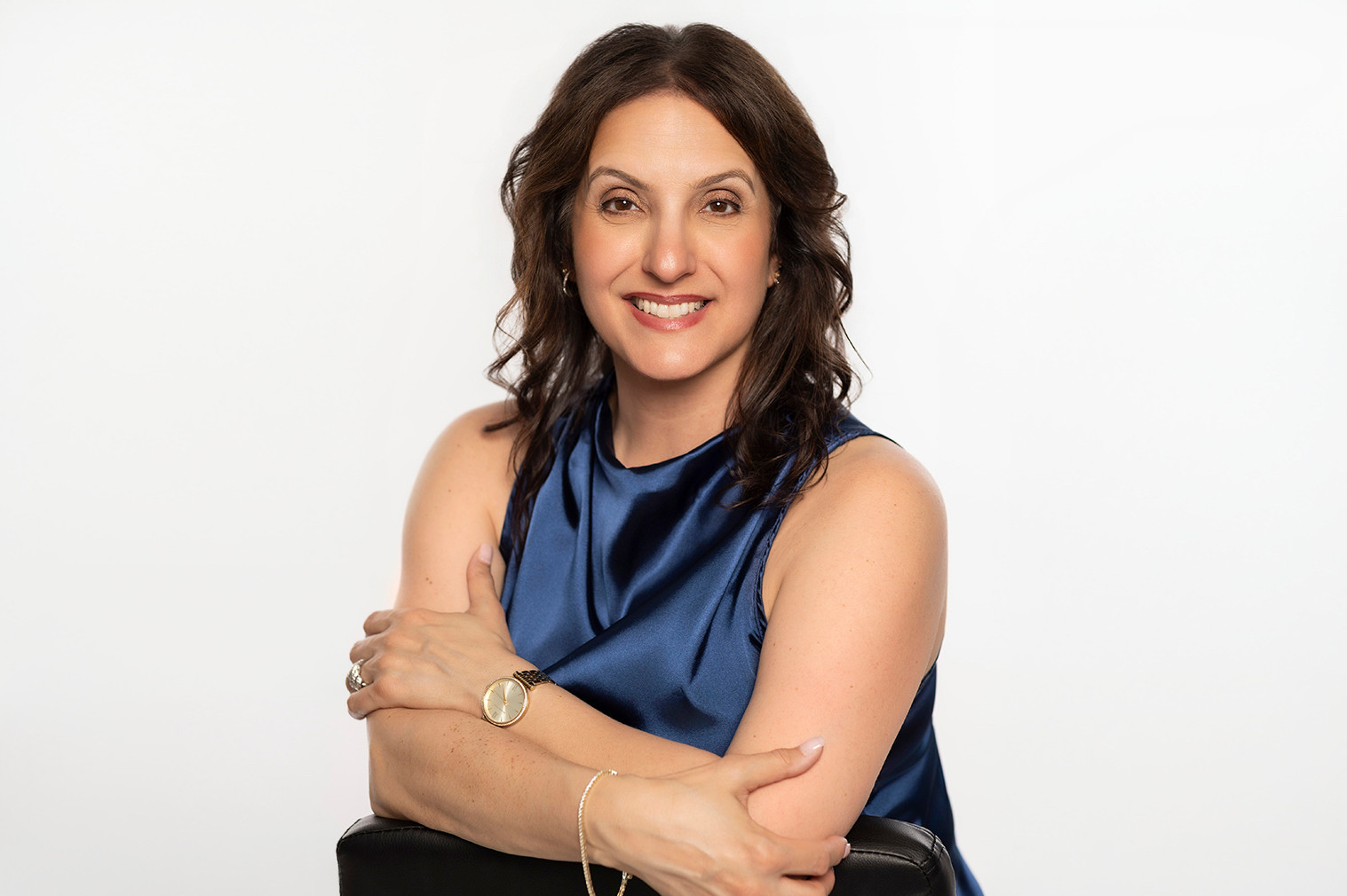 Smiling woman in a navy sleeveless top leans on a black chair against a plain white studio background.