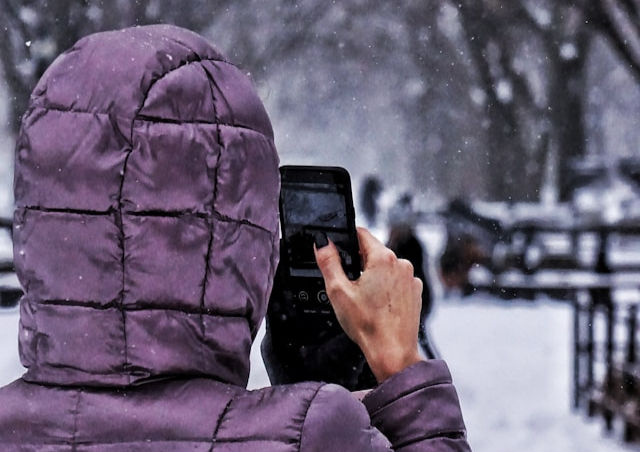 A person in a purple hooded jacket is holding a smartphone, taking a photo in a snowy outdoor setting.