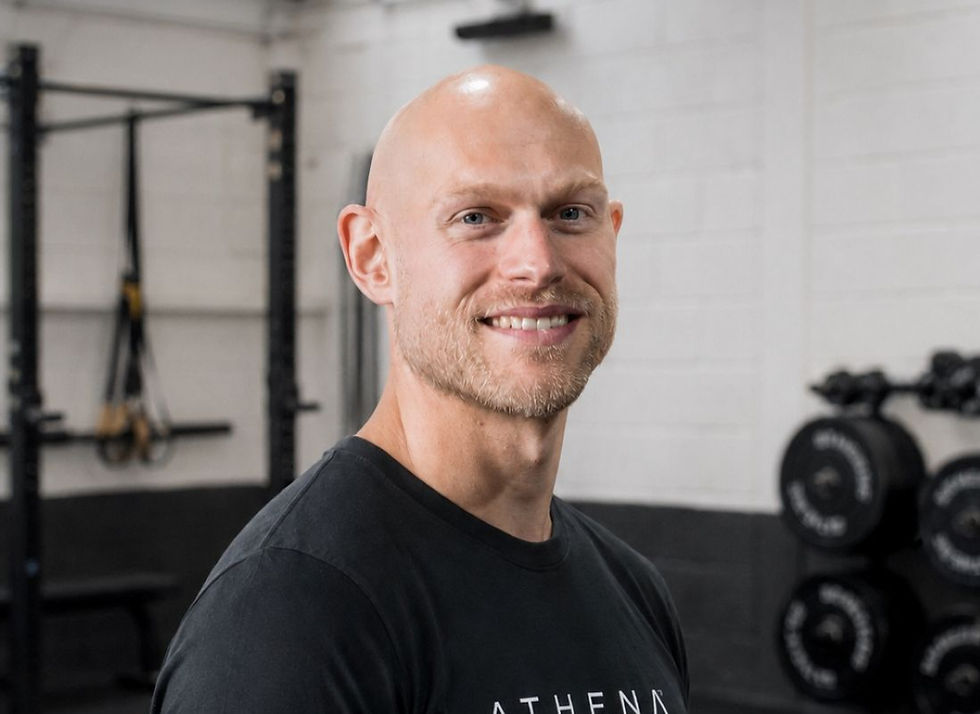 Smiling bald man in a gym, wearing a black ATHENA shirt, with racks and weight plates in the background.