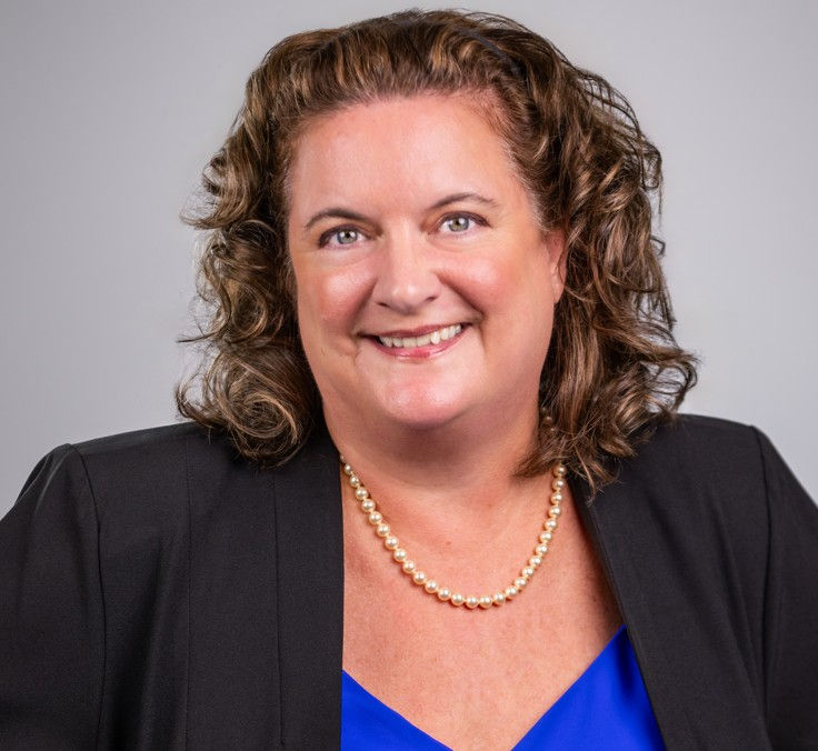 Smiling woman in a black blazer and blue top with a pearl necklace, posing for a professional headshot against a gray background.