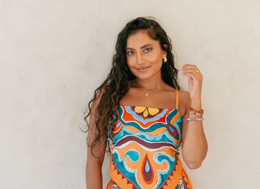 Woman smiling in colorful patterned dress, with long wavy hair, gold jewelry, neutral background. Relaxed, confident vibe.
