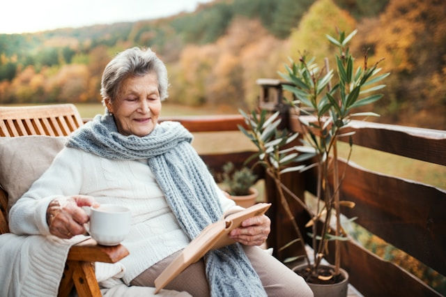 Elderly woman on a wooden porch, sipping coffee and reading, surrounded by autumn foliage. She's wrapped in a cozy gray scarf, looking content.