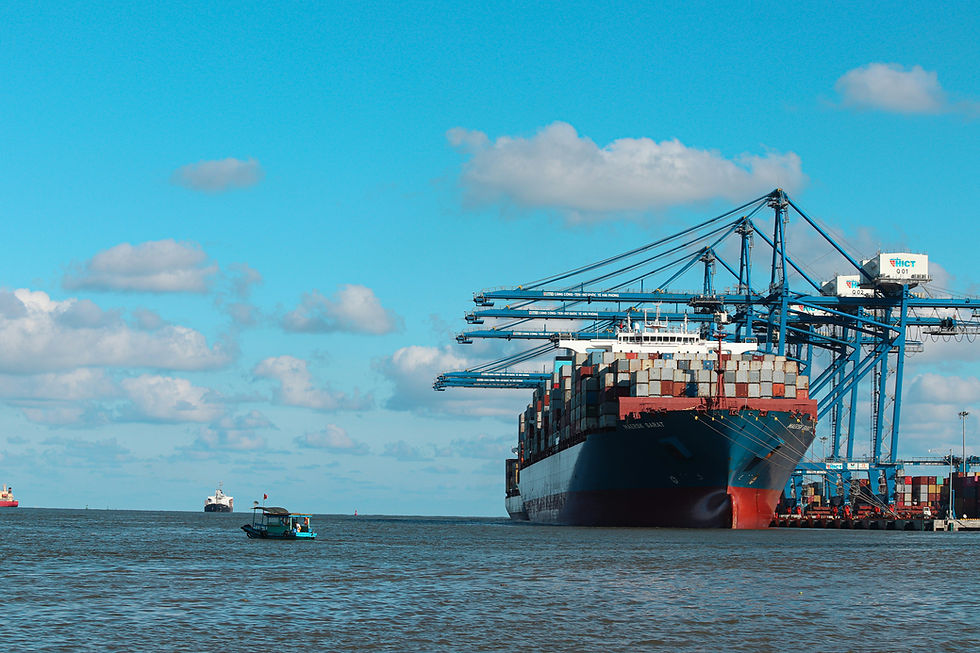 Container ship loaded with cargo sits at a port beside blue cranes, with a small boat on calm water under a bright cloudy sky.
