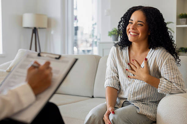 Woman with curly hair smiles, hand on chest, seated on a white sofa in a bright room. Opposite, a person holds a clipboard and pen.
