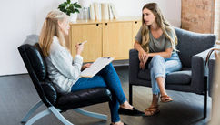 Two women in a therapy session. One takes notes while listening, sitting opposite each other in a modern room with a plant and books in the background.