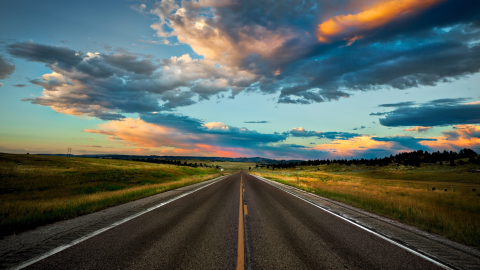 Empty two-lane road stretches through grassy plains under dramatic sunset clouds.