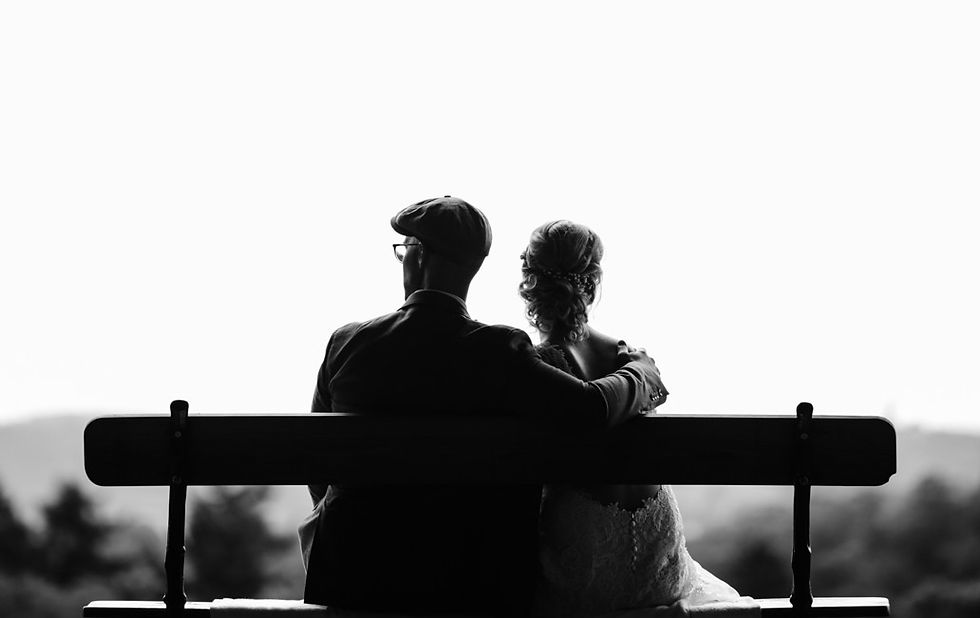 Black-and-white couple sits on a bench outdoors, man’s arm around woman, facing a soft blurred landscape in a romantic mood