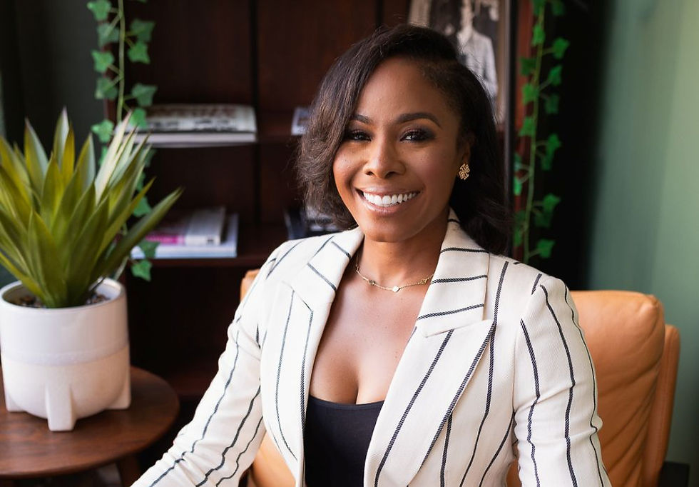 Smiling person in a white striped blazer sits in a cozy room with green walls, a potted plant, bookshelf, and framed photo behind them.