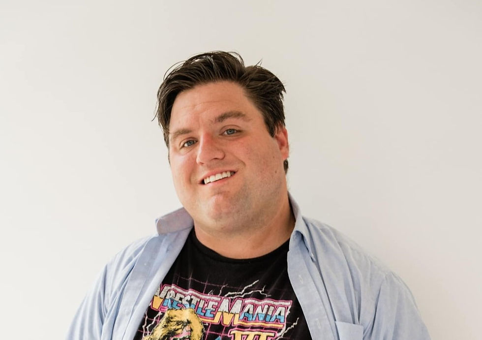 Smiling man in a WrestleMania shirt and blue shirt poses against a plain white background. Casual and relaxed mood.