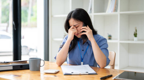 a woman sitting at a desk in a professional setting. She appears to be on a phone call and is visibly stressed or overwhelmed, as she is pinching the bridge of her nose.