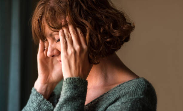 Woman in a green sweater holds her temples with eyes closed, looking stressed or in pain against a neutral indoor background