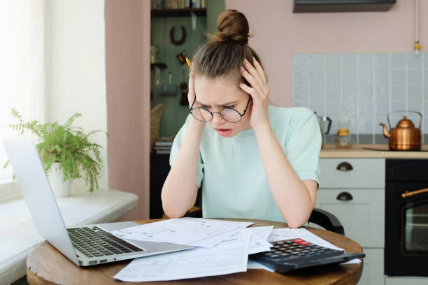 woman pressing hands to temples in helpless gesture while looking at terrifying sums and debts in her bills.
