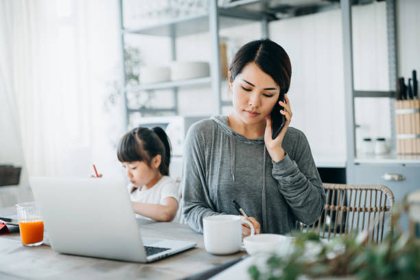 mother working from home on a laptop and talking on the phone while little daughter is studying from home.