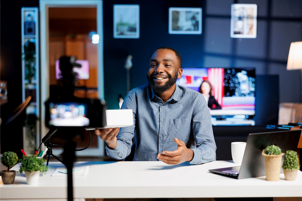 Man smiling at a desk, holding a white box toward a camera, with laptop, plants, and coffee. A TV is on in a cozy, well-lit room.