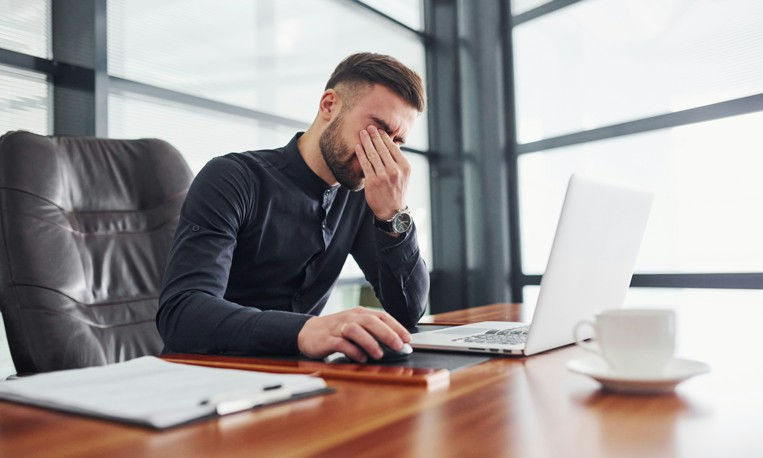 Man in black shirt at desk, rubbing his eyes, appearing stressed. Laptop, notepad, and coffee cup on wooden table. Bright office.