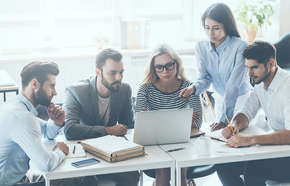 Five coworkers in a bright office huddle around a laptop, with one woman pointing at the screen while others discuss notes and papers.