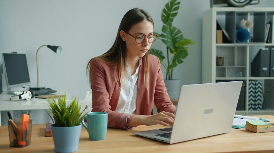 A young woman with long hair and glasses sits at a wooden desk, focused on typing on her laptop. She wears a dusty pink blazer over a white shirt. A blue mug, small plant, and office supplies sit nearby in a bright office.