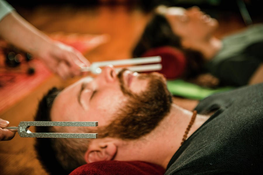 Man lies with eyes closed as tuning forks are held near his face in a calming sound-healing session indoors.