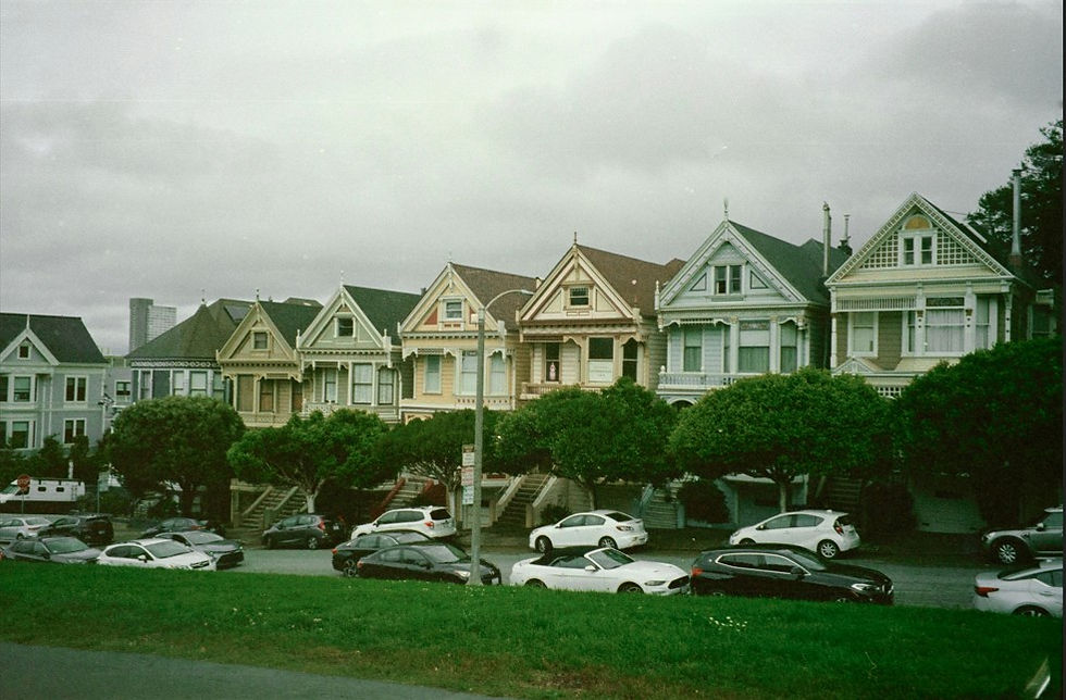 Row of colorful Victorian houses on a hilly street with parked cars and cloudy sky, calm city neighborhood.