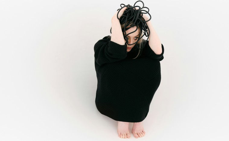 A person in a black outfit crouches on a white background, holding black cables over their head, creating a contemplative, surreal mood.