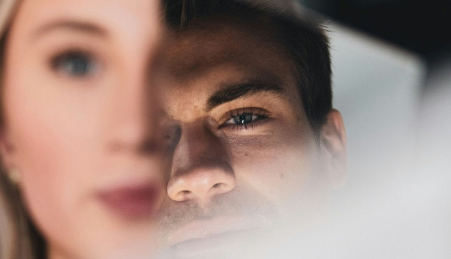 Close-up of a man peering past a blurred female face in the foreground, with a moody soft-focus dark background.
