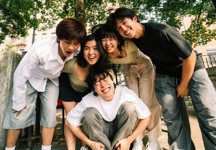 Five friends laughing together outside under trees, leaning in for a group hug. Casual clothing, joyful mood, summer day.
