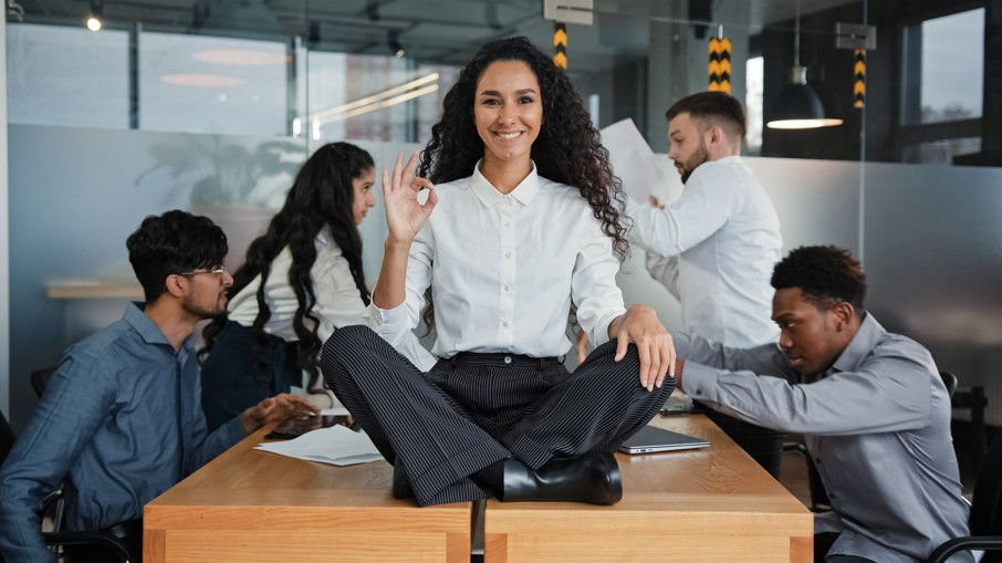 A smiling woman sits cross-legged on an office desk, making an "okay" hand sign. She maintains a calm, composed expression while surrounded by chaotic coworkers arguing and tossing papers in a busy, modern office environment.