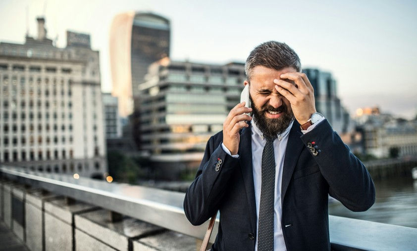 Stressed bearded businessman in a dark suit talks on a phone on a bridge, rubbing his forehead with city buildings behind.