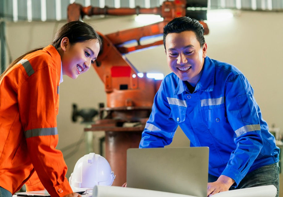 Two smiling factory workers in orange and blue uniforms lean over a laptop in a workshop, with a robotic arm behind them.
